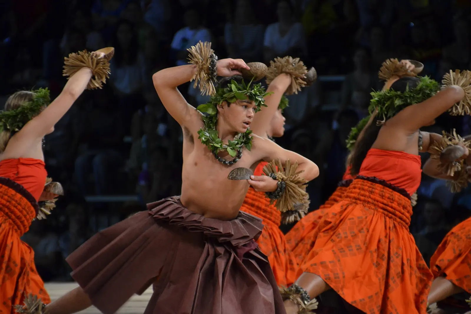 A hula kahiko dancer in a leaf crown and brown skirt mid-step on stage, with dancers in orange skirts beside him