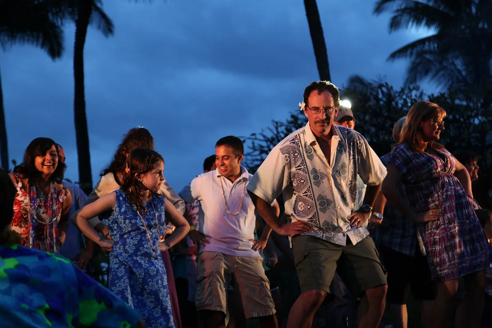 Guests dancing together outdoors at dusk at a luau, with palm trees and warm lights behind